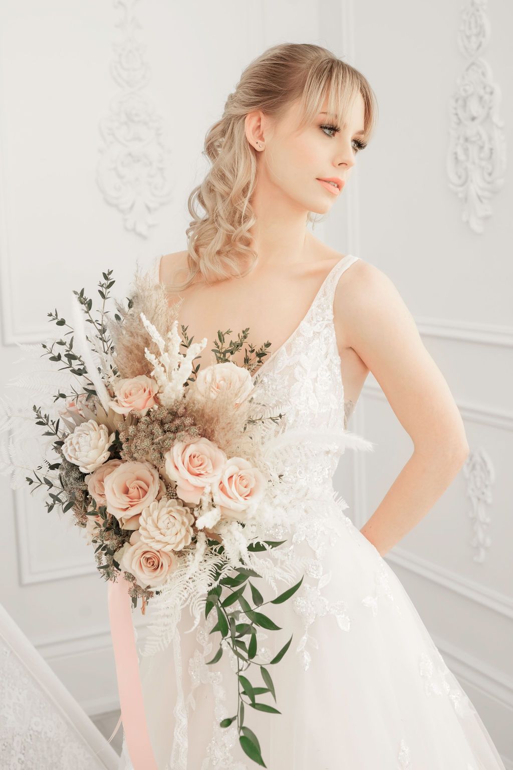 Blonde bride in a white dress holding a bouquet of pink roses, looking elegant in a bright room.