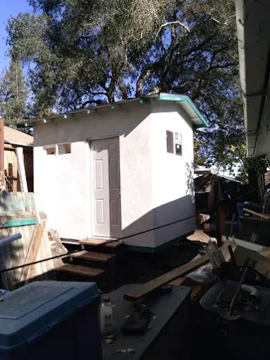 Small white shed with light blue trim.  A door and window are visible. The shed is on wooden steps.