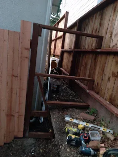 Wooden fence construction with tools, framing, and new fence panels. Outdoors, narrow space.