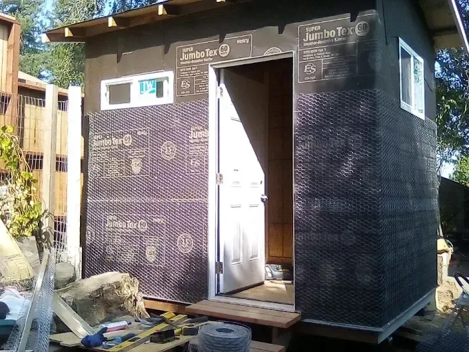 Small shed under construction, exterior with black mesh, door, and window. Sunlight streams from the open doorway.