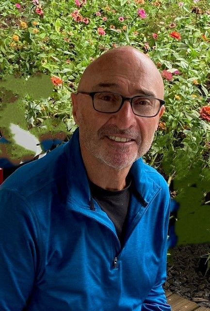 A man wearing glasses and a blue jacket is smiling in front of flowers.