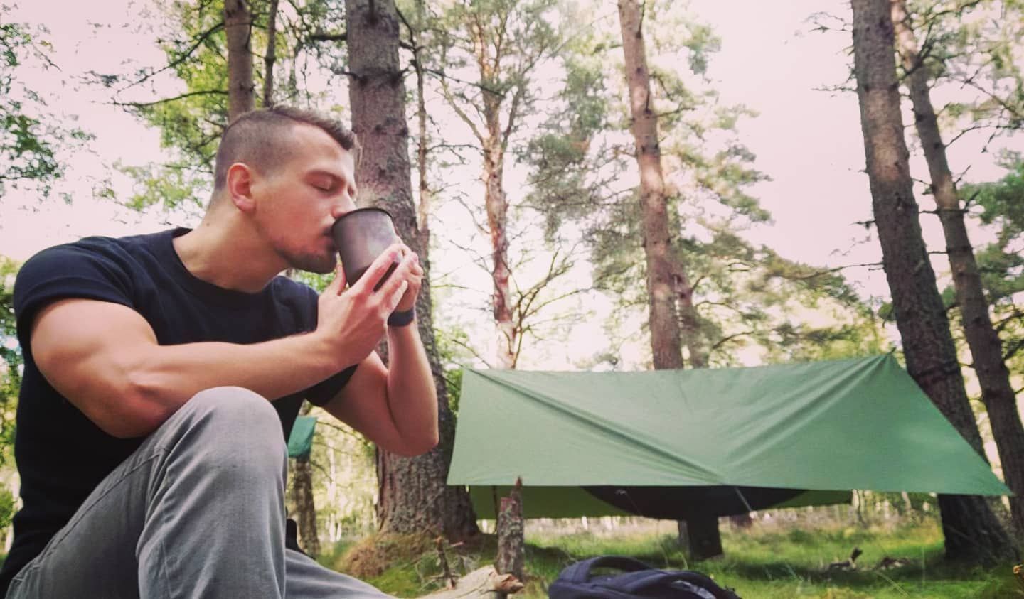 Man drinking from a mug outdoors, forest background. Green tarp shelter.