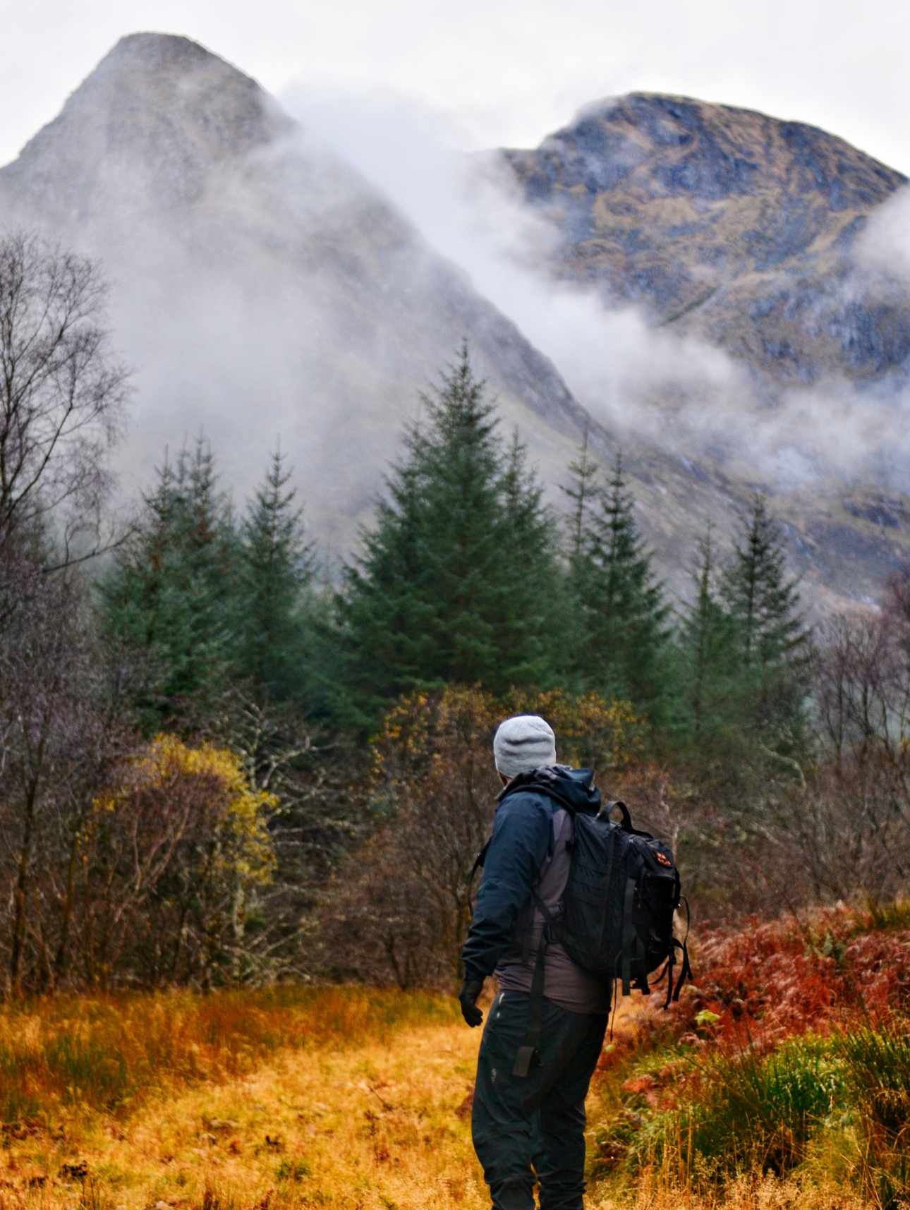 Hiker in outdoor gear looking at a mountain partially obscured by clouds; autumn forest.