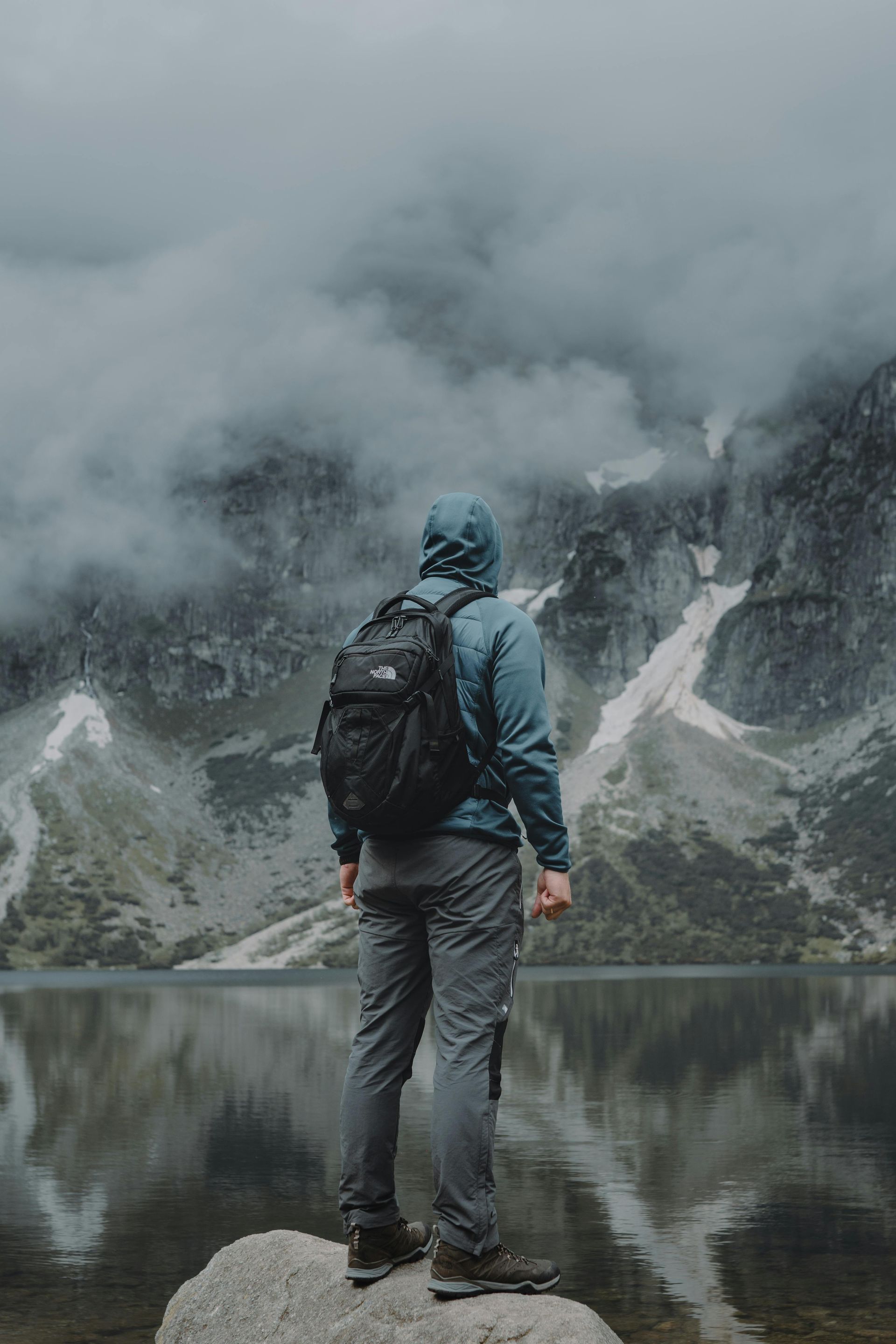 Person with backpack standing on a rock, facing mountains, cloudy sky, lake in foreground.