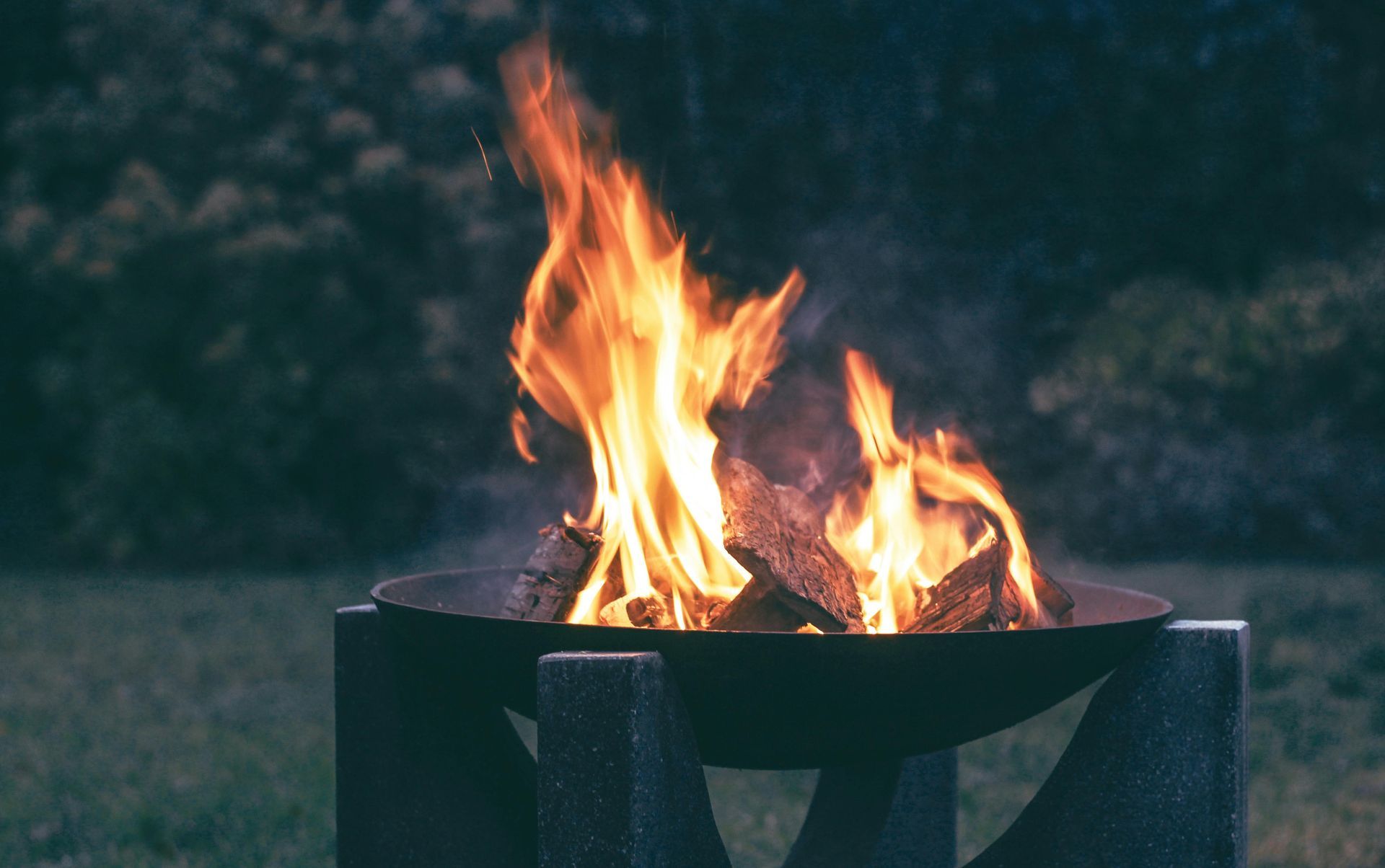 Campfire burning brightly in a dark metal bowl. Flames are orange and yellow, set outdoors.