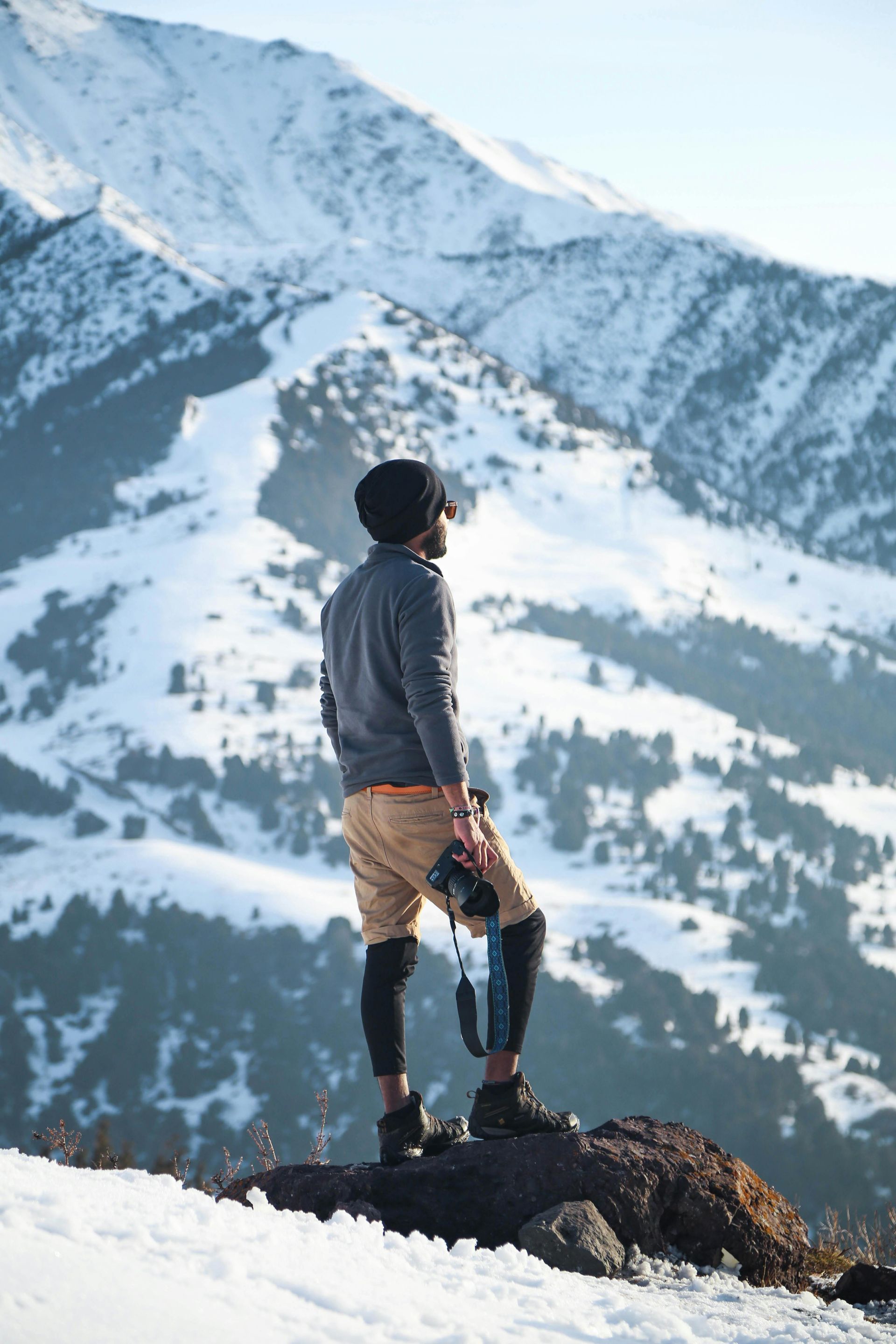 Man stands on a snowy mountain, holding a camera, looking at snow-covered peaks.