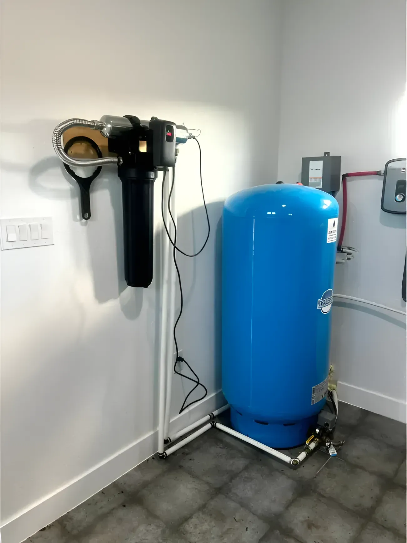 Water filtration and storage system in a corner of a room, with black filter, blue tank, and white piping.