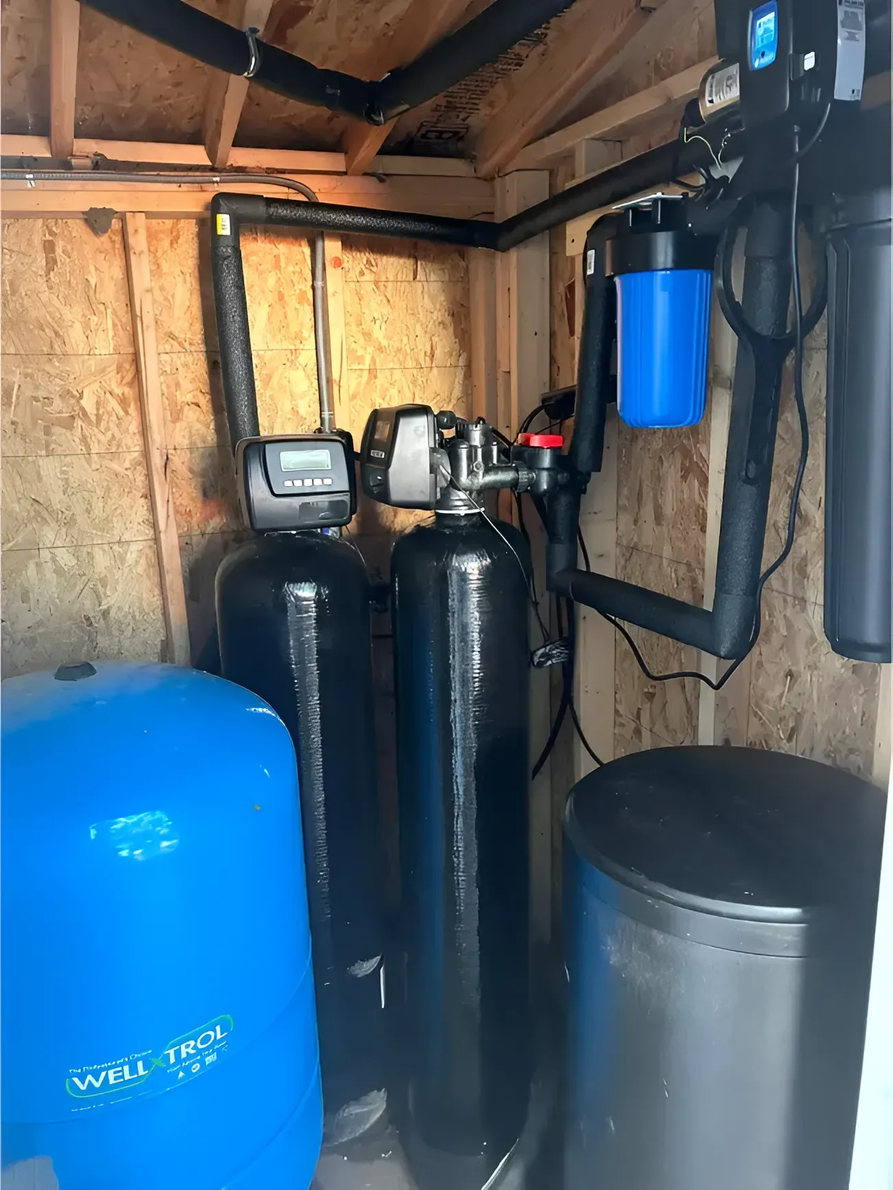 Water filtration system inside a small wooden enclosure, including tanks, filters, and pipes.