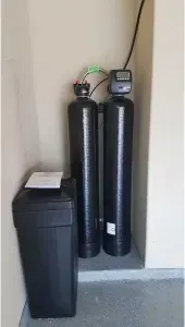 Water softener system in a garage corner: black tanks, digital display, and salt container.