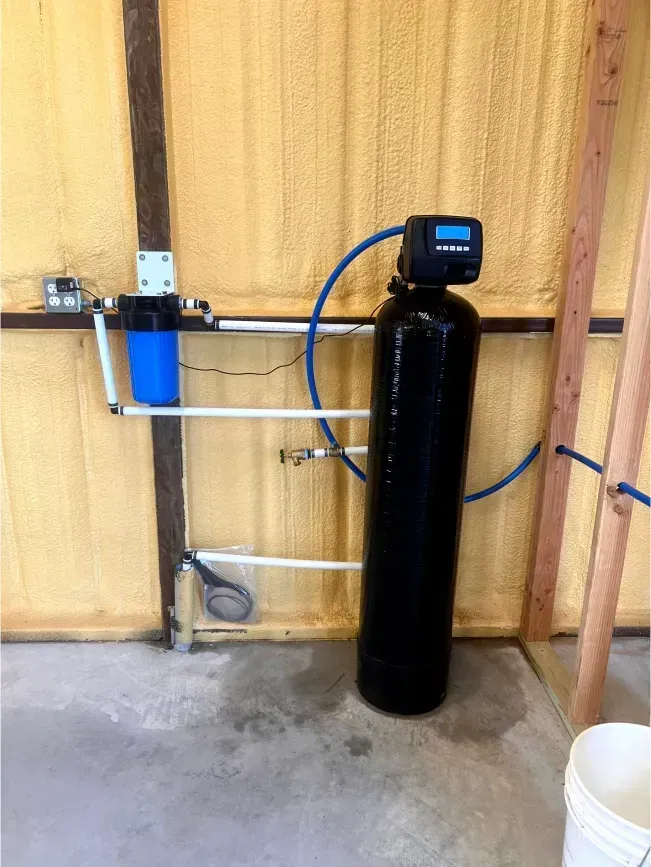 Water filtration system installed in a room with yellow insulation. Black tank, blue filter, white pipes.