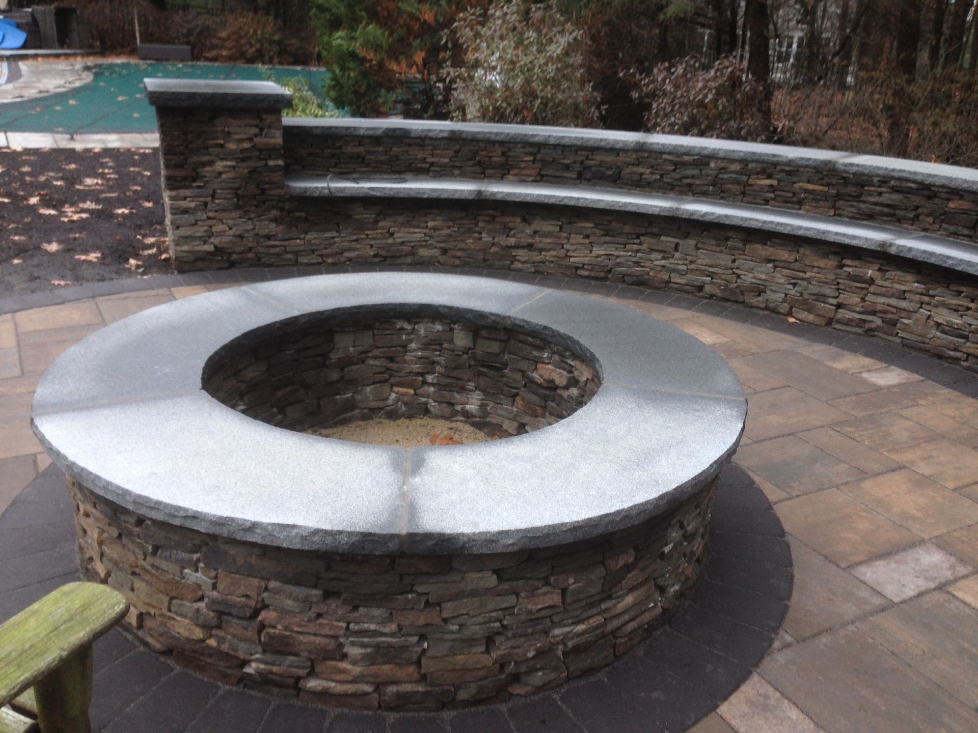 A fire pit is surrounded by a stone wall on a patio next to a pool.