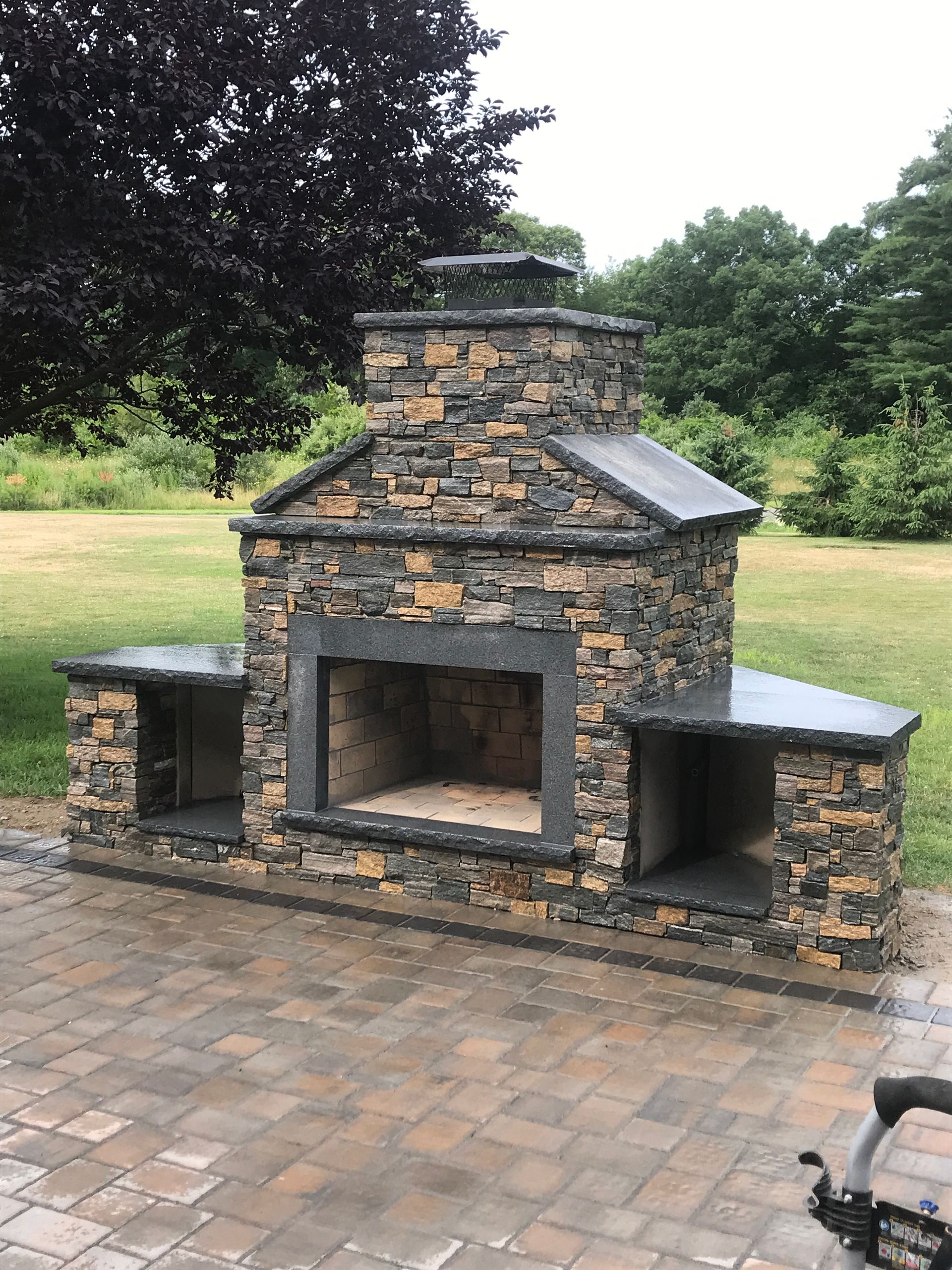 A stone fireplace is sitting on a brick patio next to a tree.