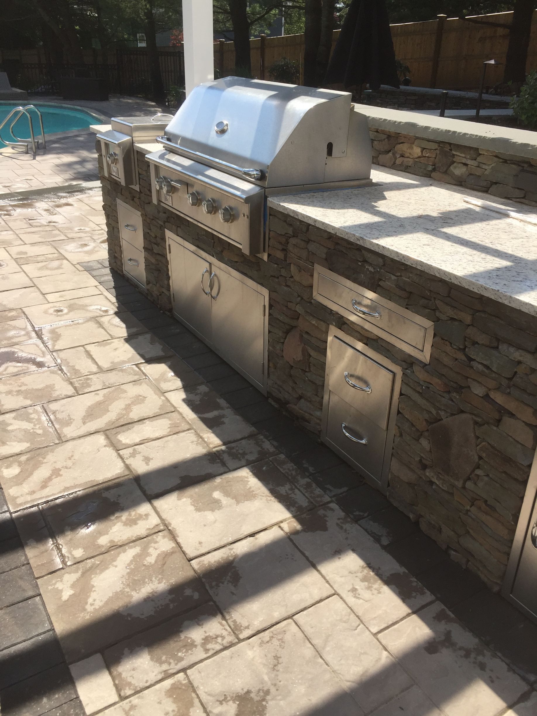 A stainless steel grill is sitting on top of a stone counter next to a pool.