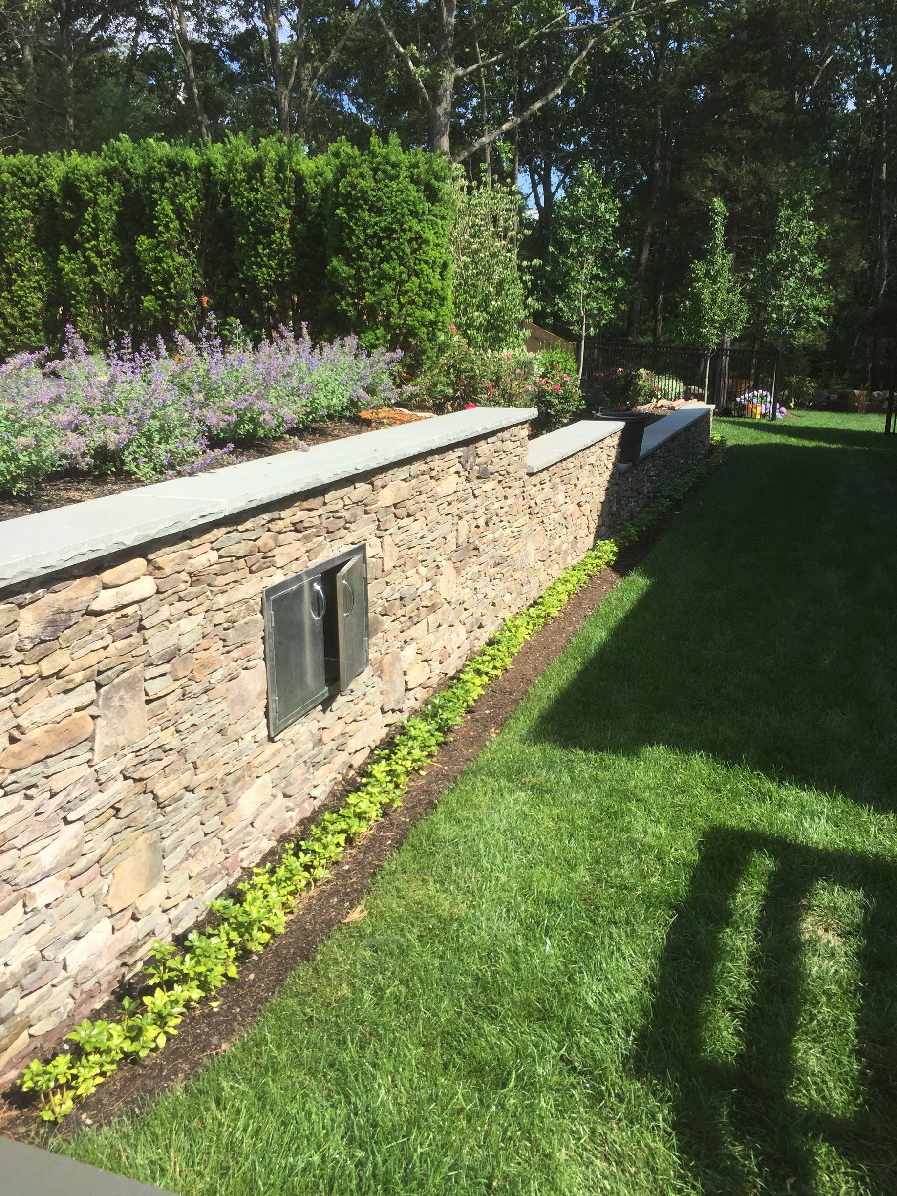 A stone wall surrounds a lush green lawn in a backyard.