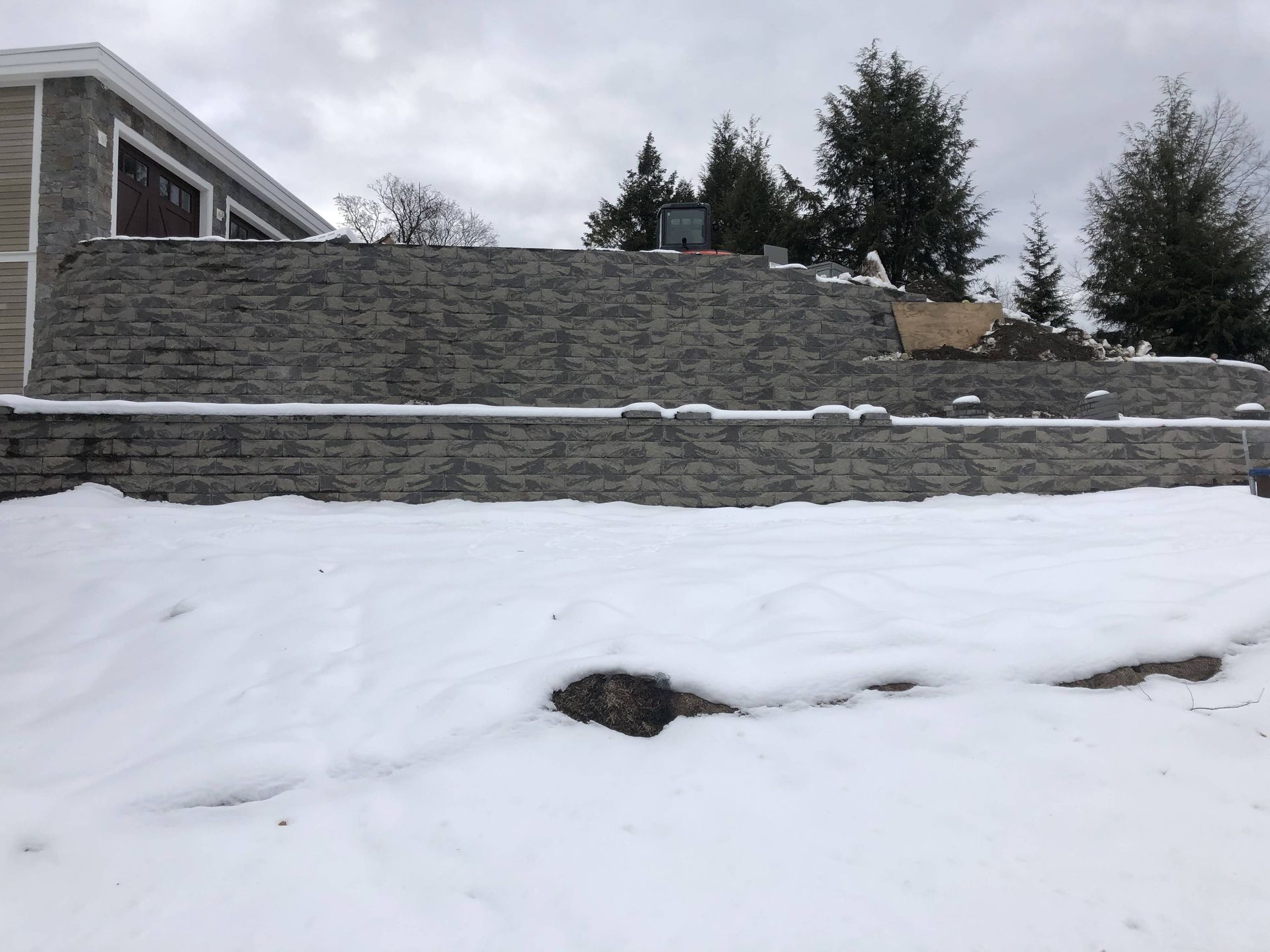A large stone wall is surrounded by snow in front of a house.