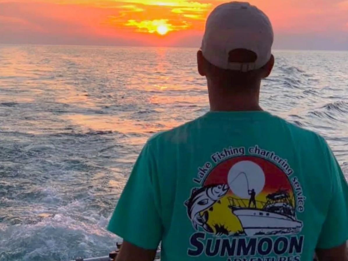 A man wearing a sunmoon shirt looks at the sunset over the ocean