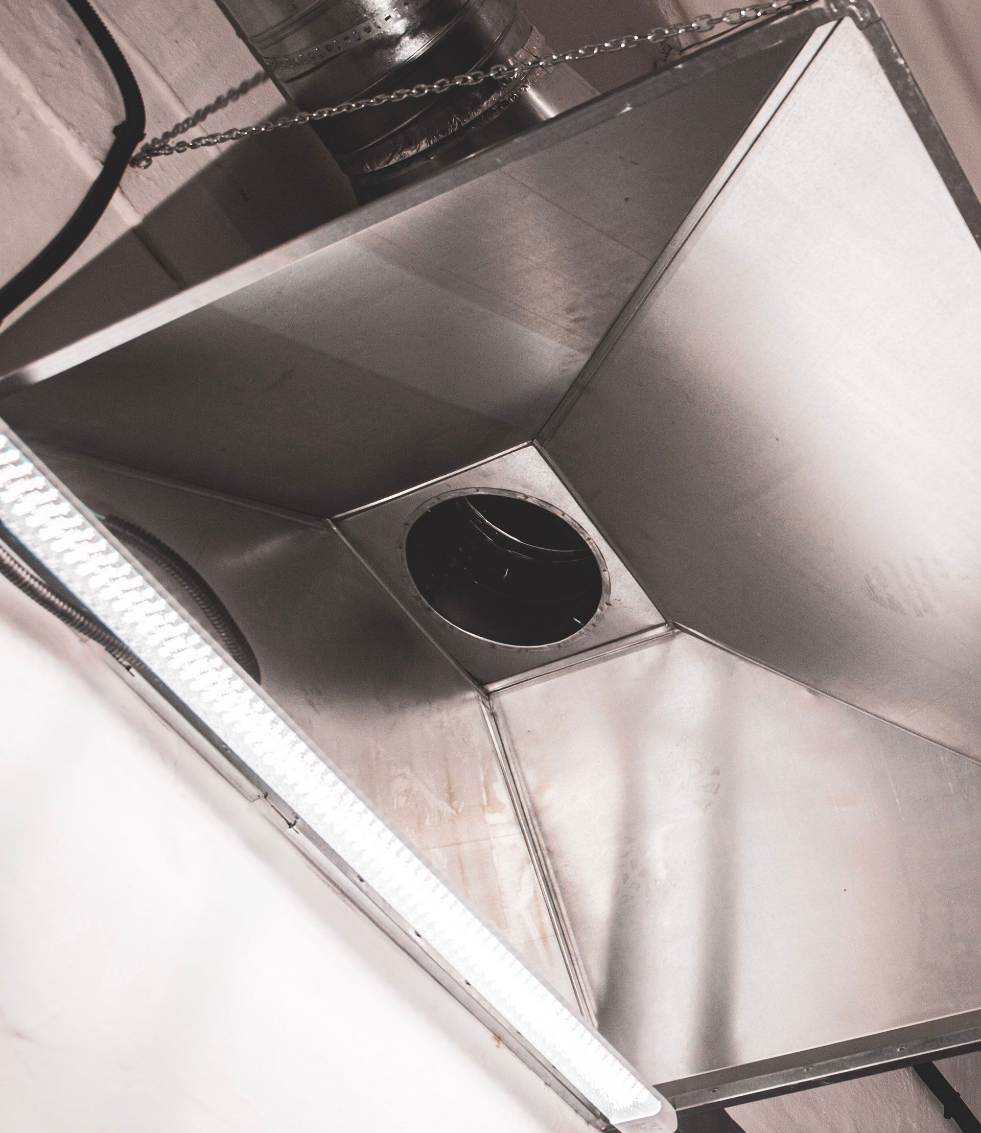 A metal, square-shaped ventilation hood with a circular exhaust port suspended by chains from a ceiling.