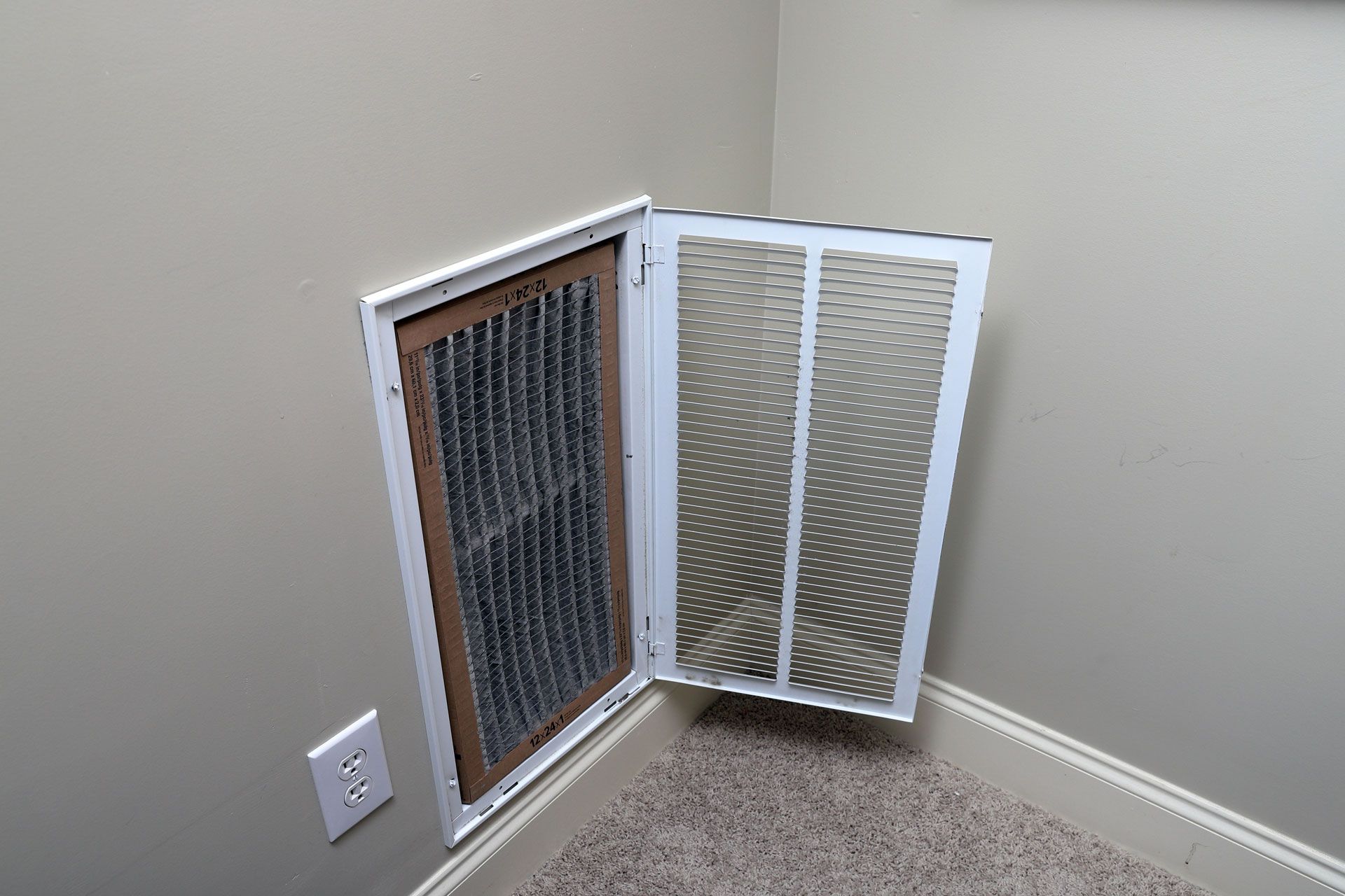 A white wall-mounted HVAC return vent with its cover open, revealing a rectangular air filter inside.
