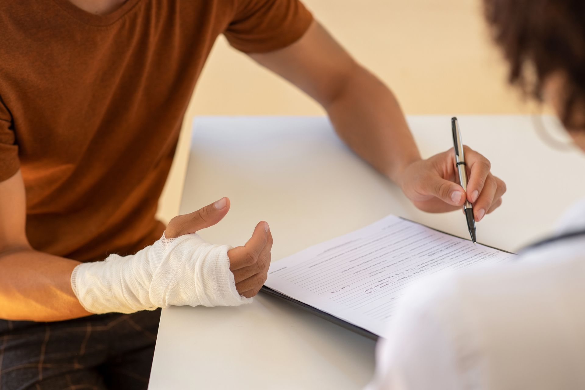 Person with a bandaged hand talking with a doctor at a desk. The doctor is holding a pen and a clipboard.