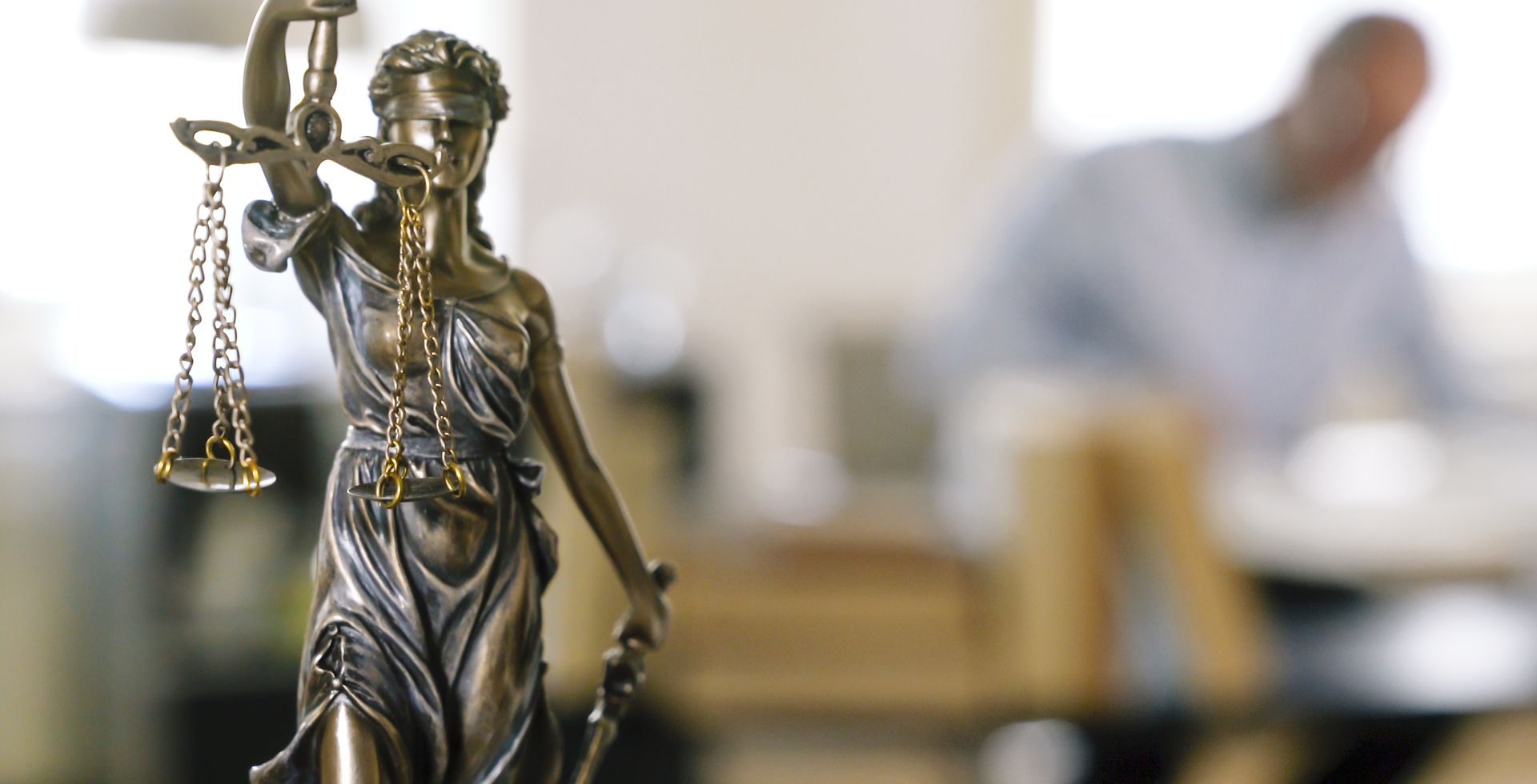 Lady Justice statue with scales in law office setting, blurred legal professional in background.