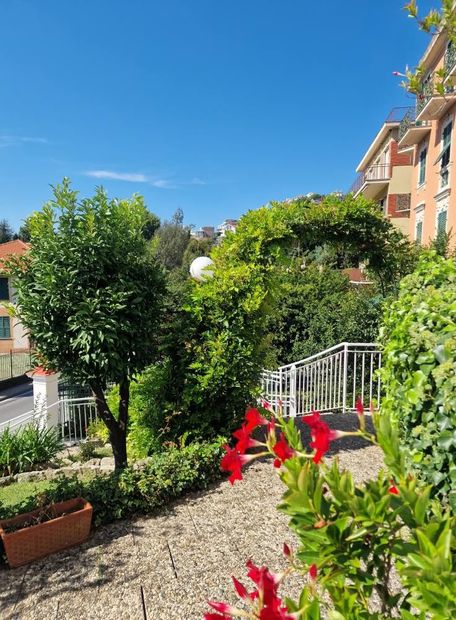 Scena di giardino con fiori rossi, alberi, sentiero in pietra, scale e un edificio sotto un cielo azzurro.