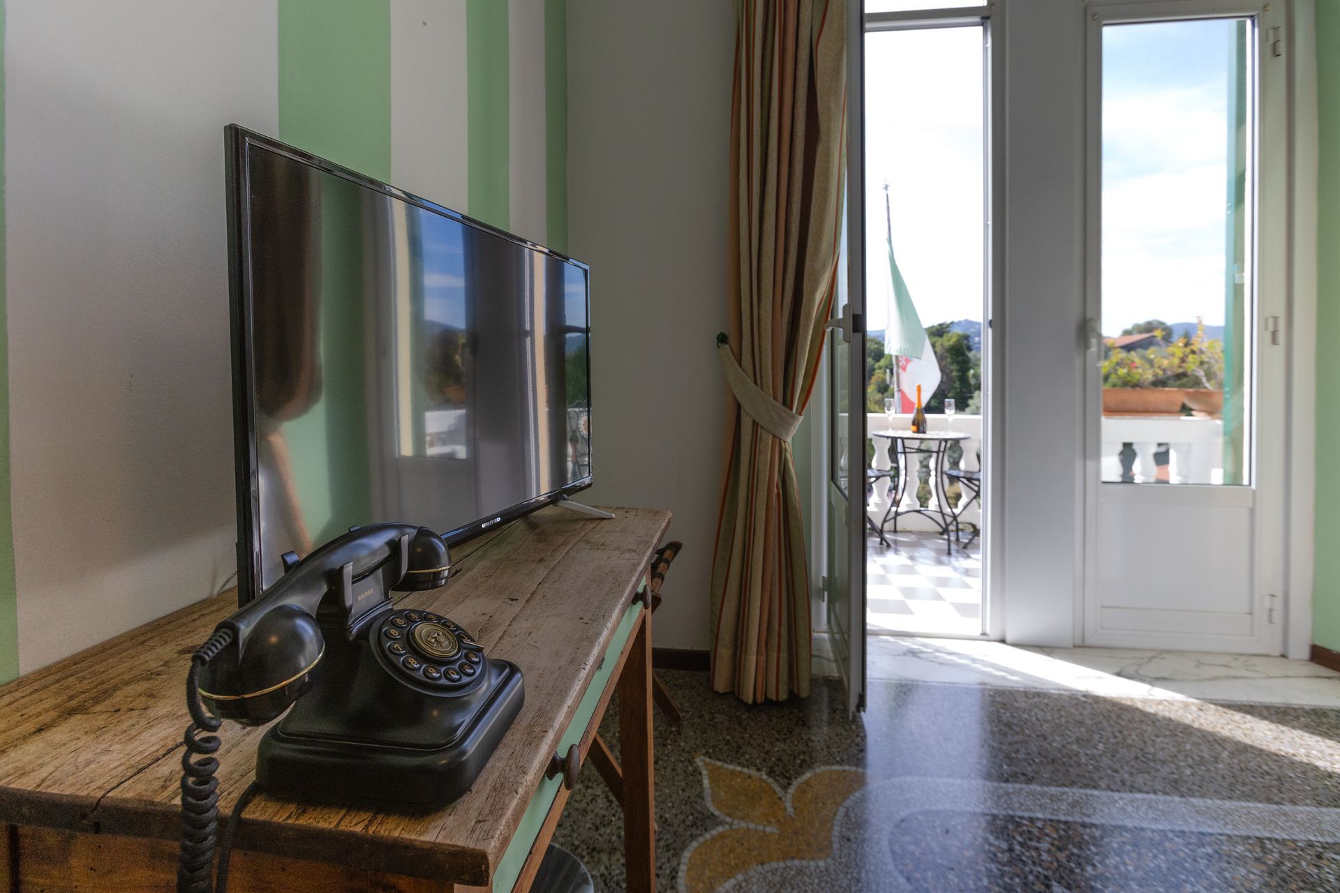 Una stanza con TV, telefono antico e una porta aperta su un balcone con la bandiera italiana.