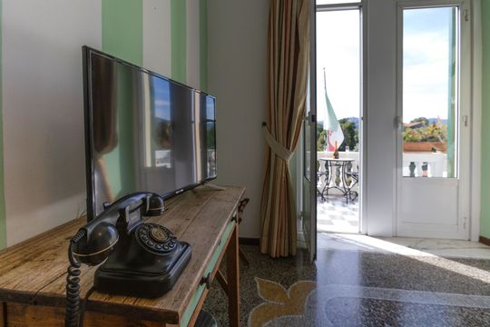 Una stanza con TV, telefono antico e una porta aperta su un balcone con la bandiera italiana.