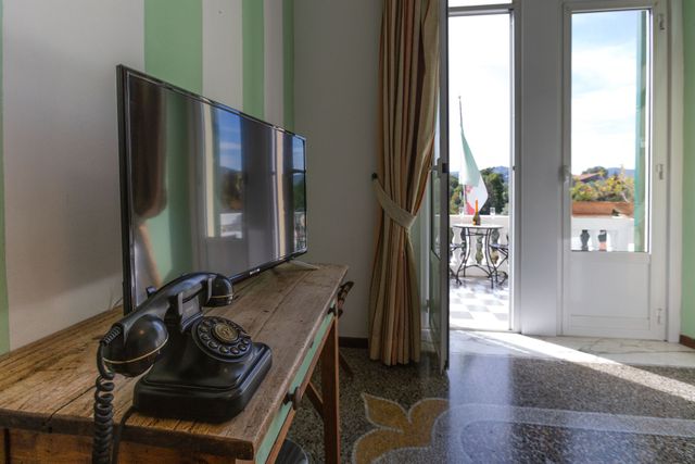 Una stanza con TV, telefono antico e una porta aperta su un balcone con la bandiera italiana.