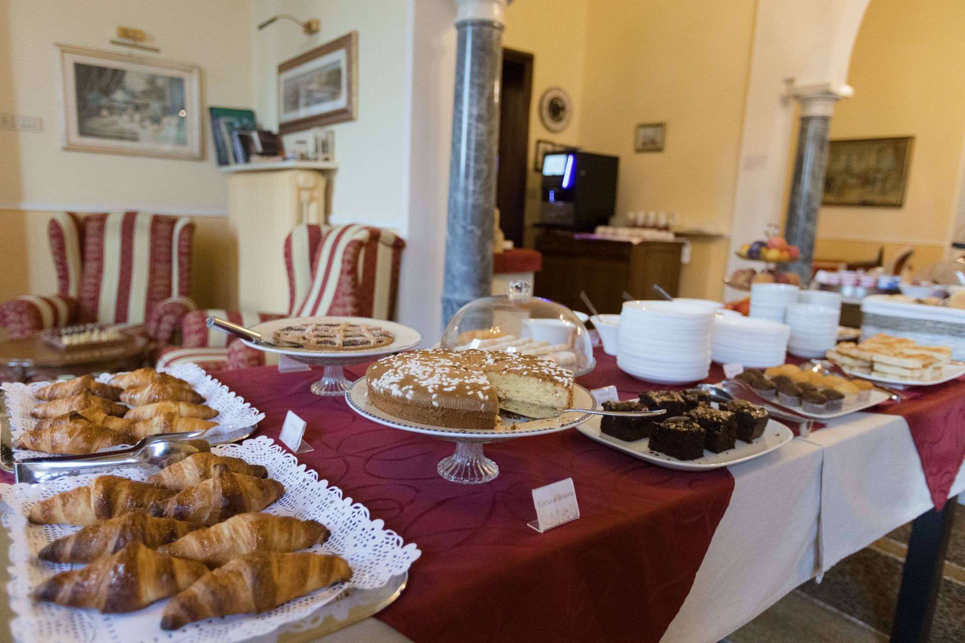 Tavolo per la colazione a buffet con pasticcini, torte e caffè, allestito in una sala da pranzo con colonne classiche.