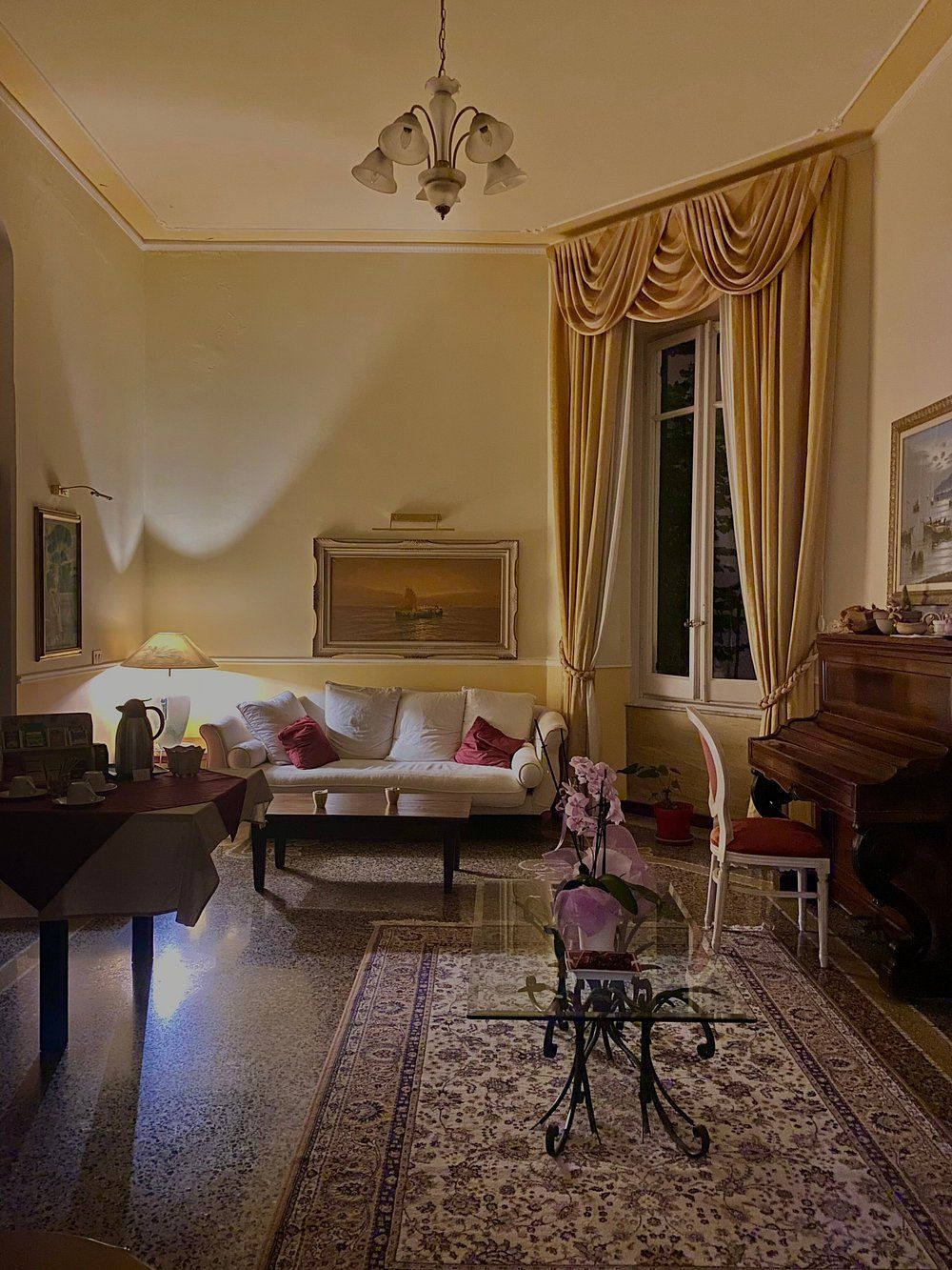 Accogliente soggiorno con pareti color crema, tappeto a fantasia, divano e finestra con tende drappeggiate. Pianoforte e tavolo a vista.