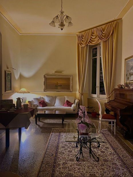 Accogliente soggiorno con pareti color crema, tappeto a fantasia, divano e finestra con tende drappeggiate. Pianoforte e tavolo a vista.