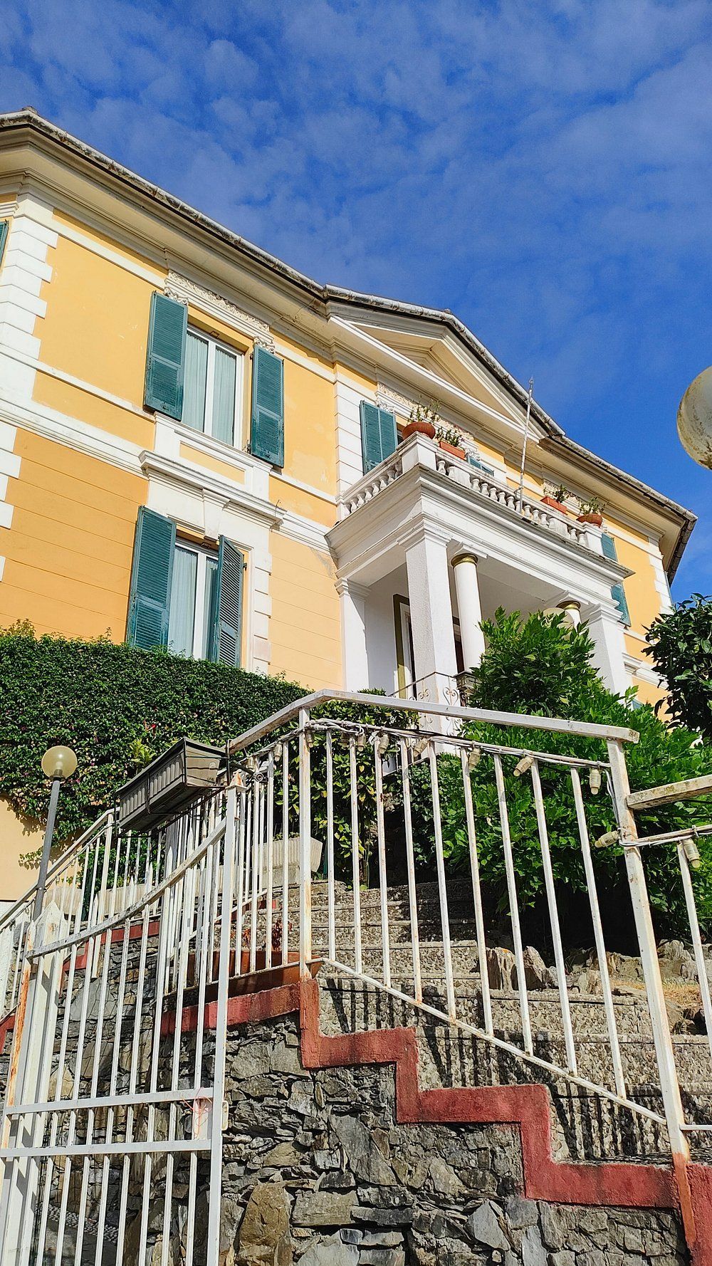 Edificio giallo con persiane verdi, balcone bianco e scale con ringhiera bianca. Cielo azzurro sullo sfondo.