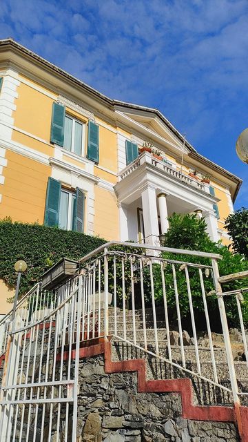 Edificio giallo con persiane verdi, balcone bianco e scale con ringhiera bianca. Cielo azzurro sullo sfondo.