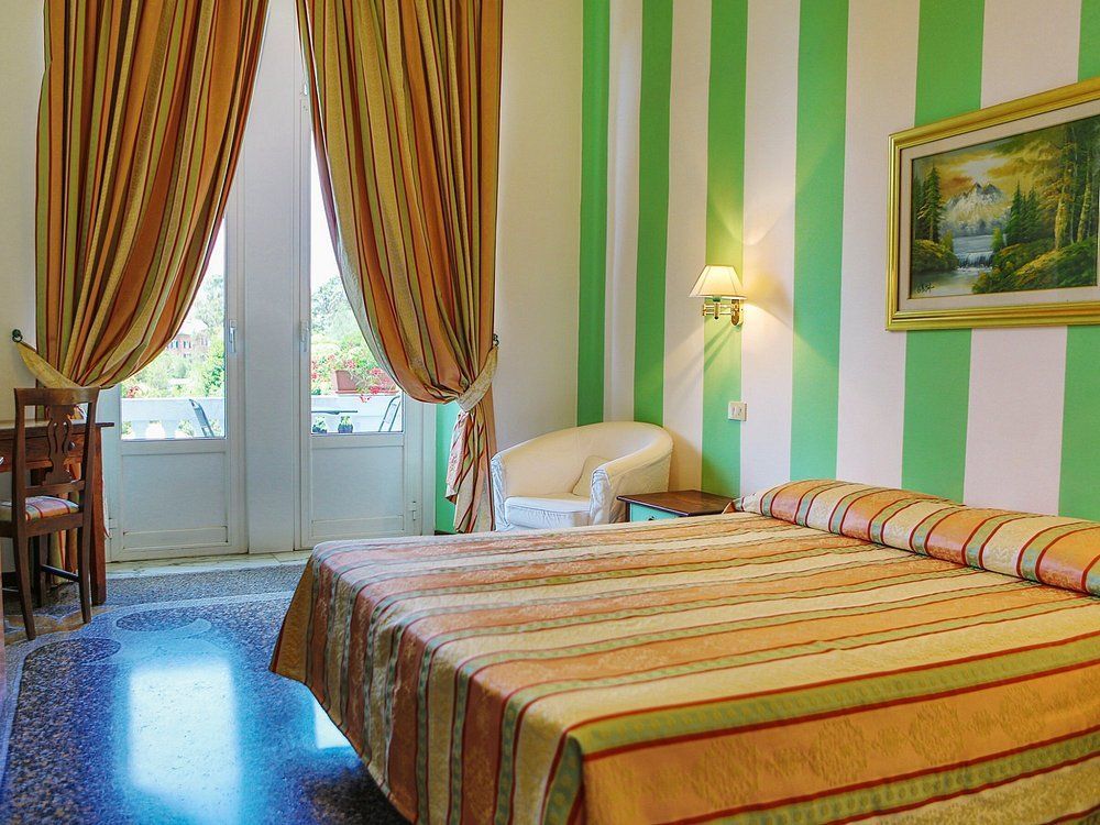 Camera da letto con parete a strisce verdi e bianche, letto con copriletto a righe, porte-finestre aperte e quadro.