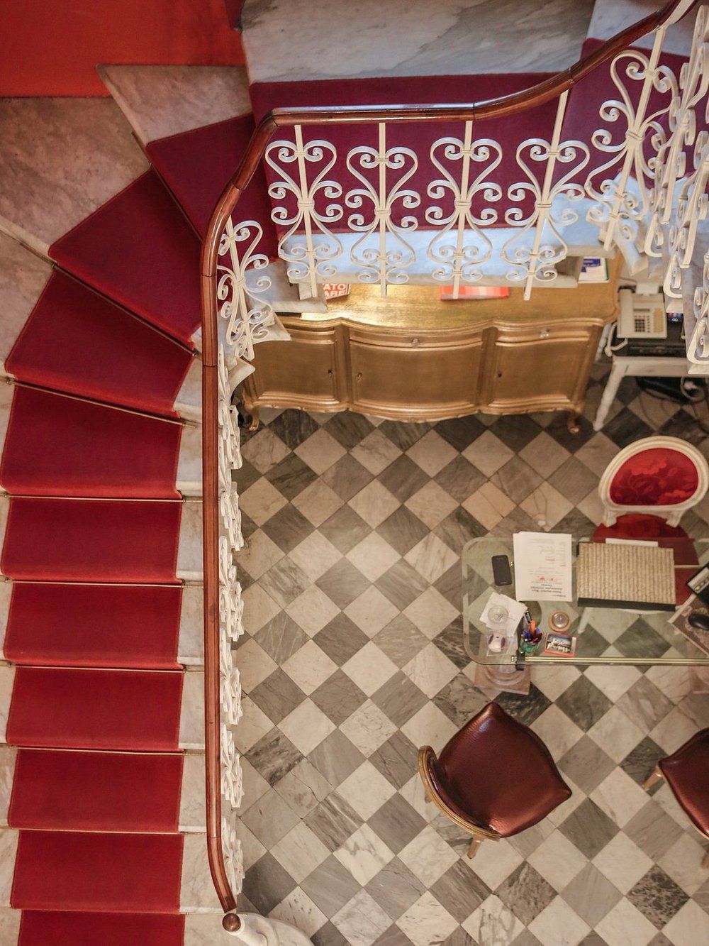 Scala con moquette rossa e ringhiera bianca decorativa. Pavimento in piastrelle a scacchi, sedie rosse.