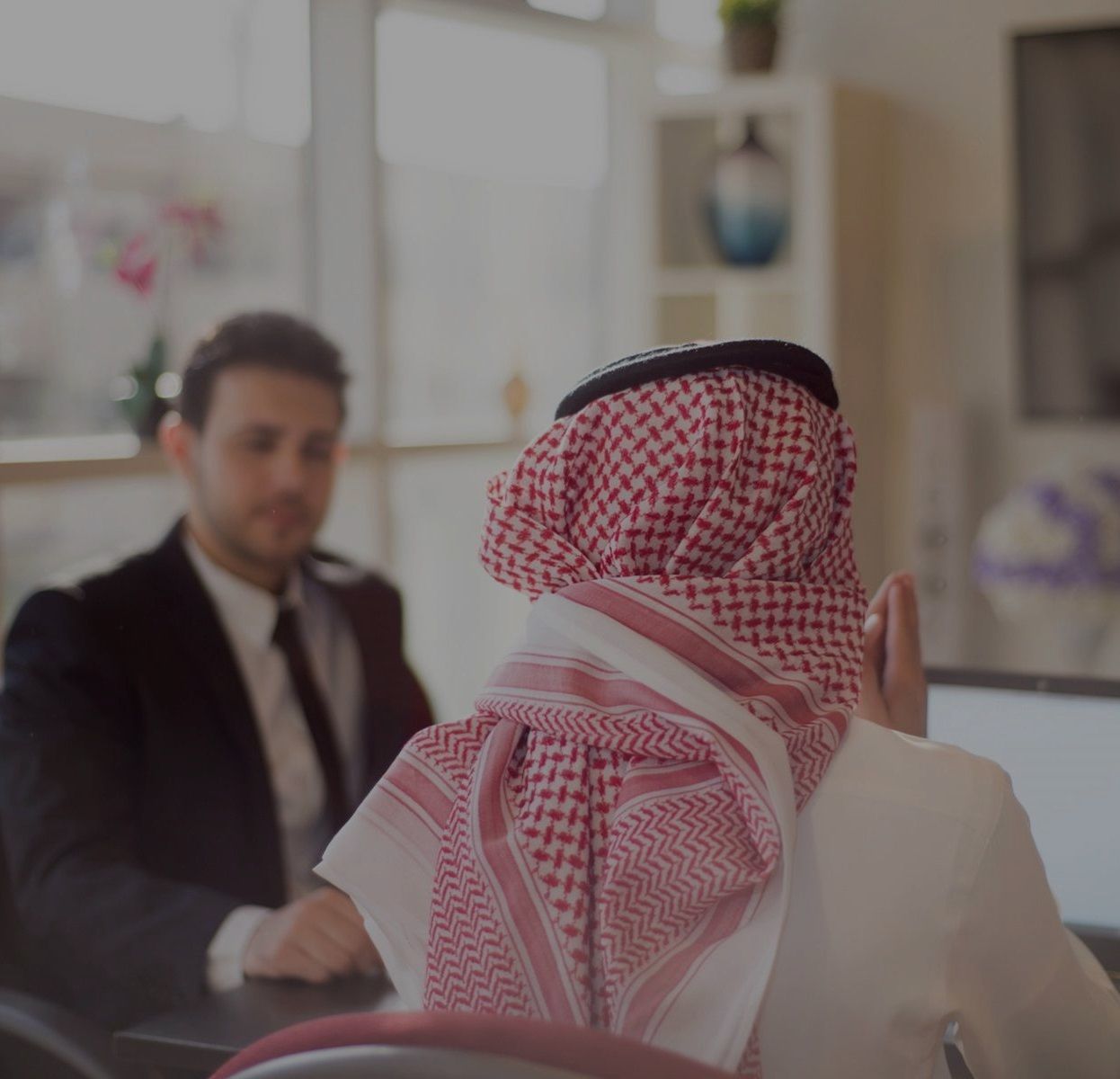 Man in traditional headwear gestures, facing another man in a suit at a table.