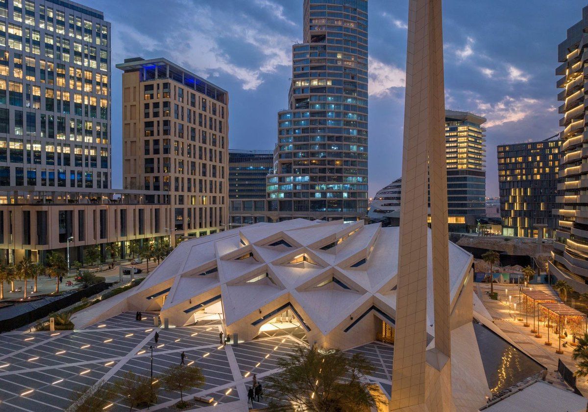 Modern mosque with geometric roof in a city square, surrounded by high-rise buildings at dusk.