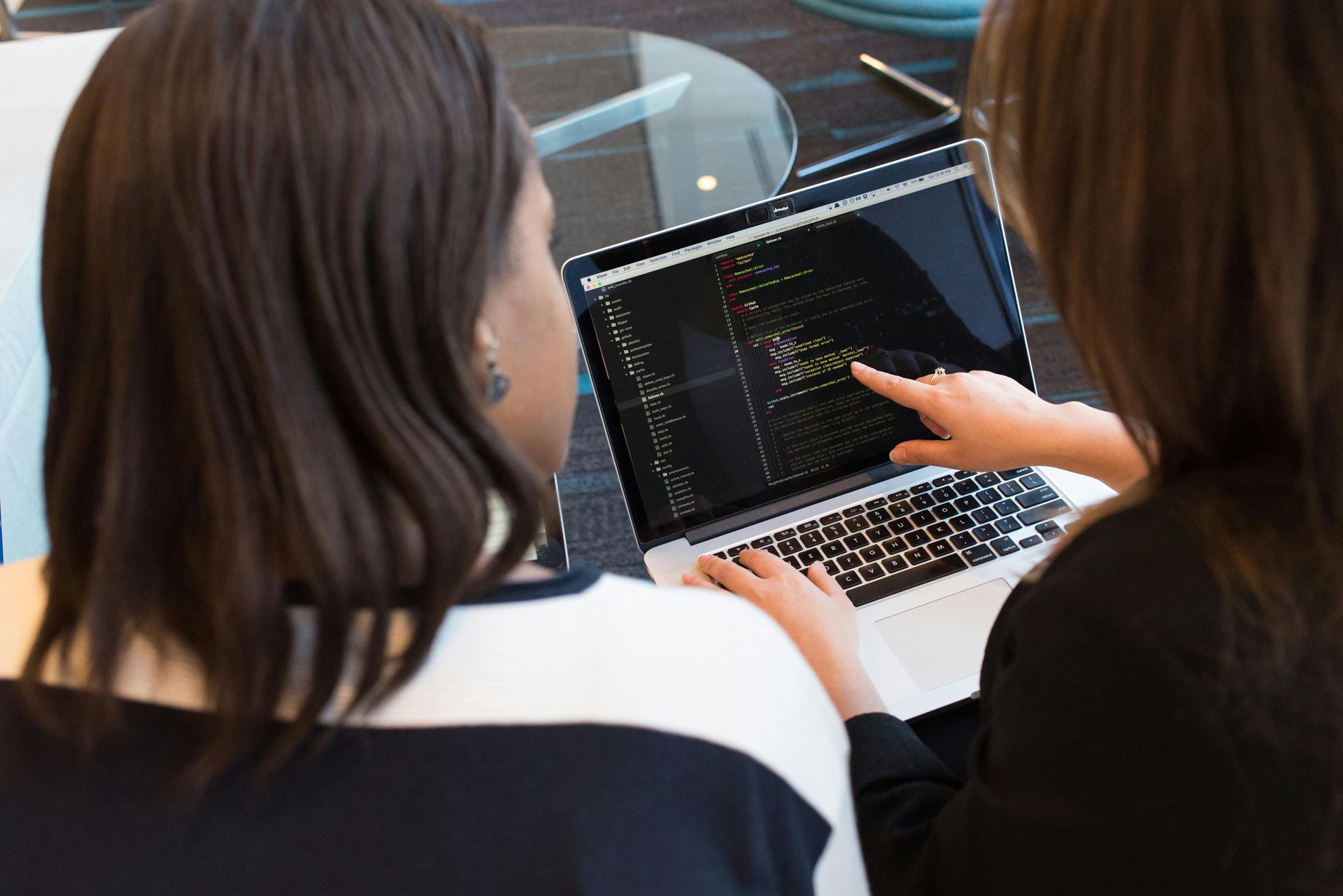 Two women looking at a laptop screen, one pointing at code, in an office setting.