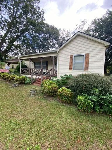 Tan house with a porch and brown shutters, surrounded by green shrubs and grass.