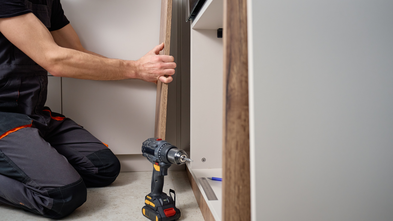 A worker kneels on the floor, holding a wooden panel next to a cabinet frame with a cordless drill resting nearby.