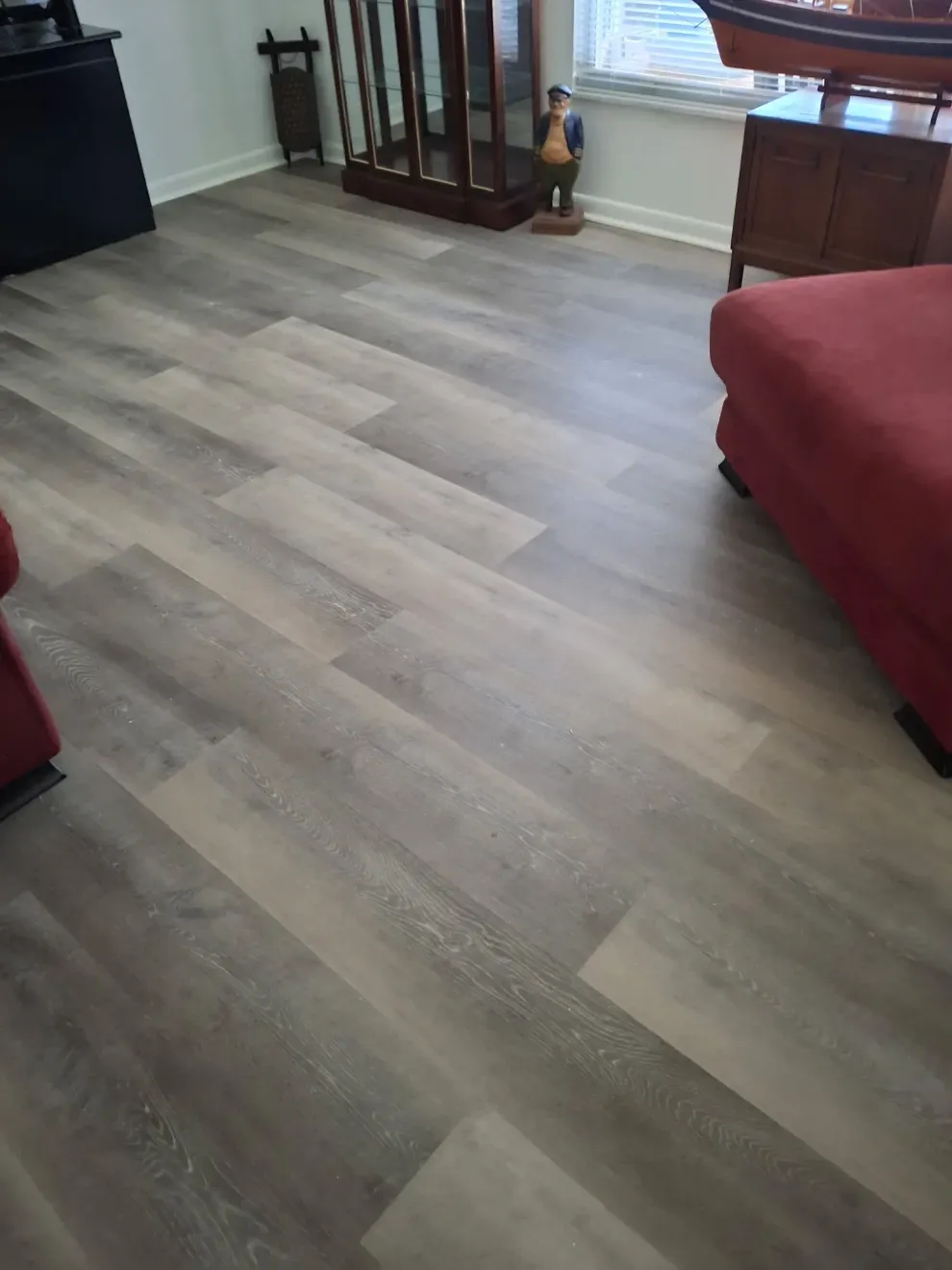 A living room featuring gray, wood-look vinyl plank flooring, with red furniture and a display cabinet visible.