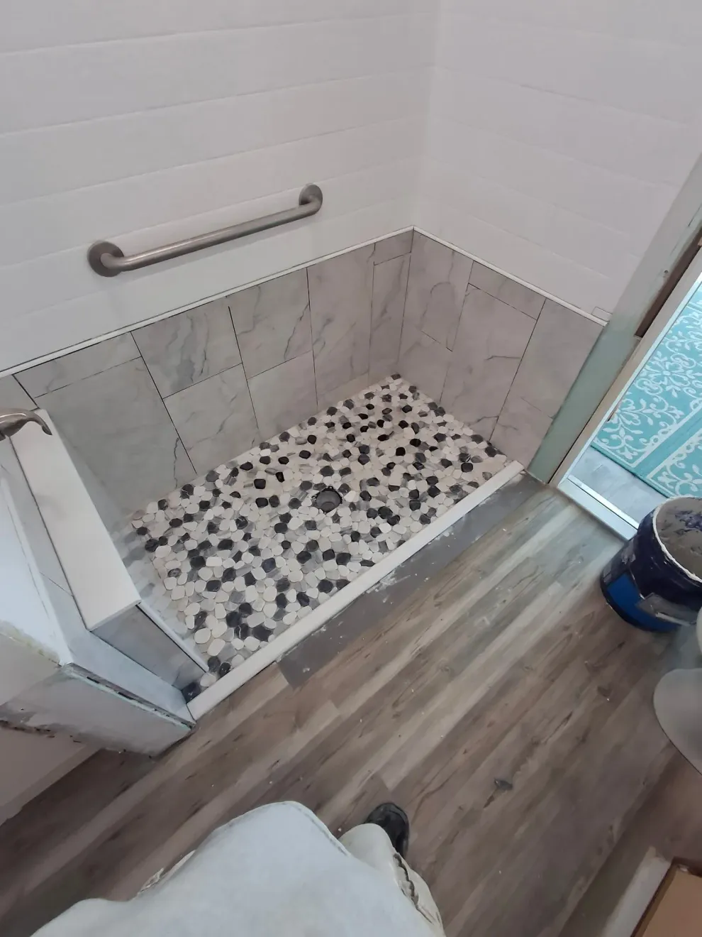 A walk-in shower with light grey tiled walls, a black and white pebble floor, and a horizontal metal grab bar.