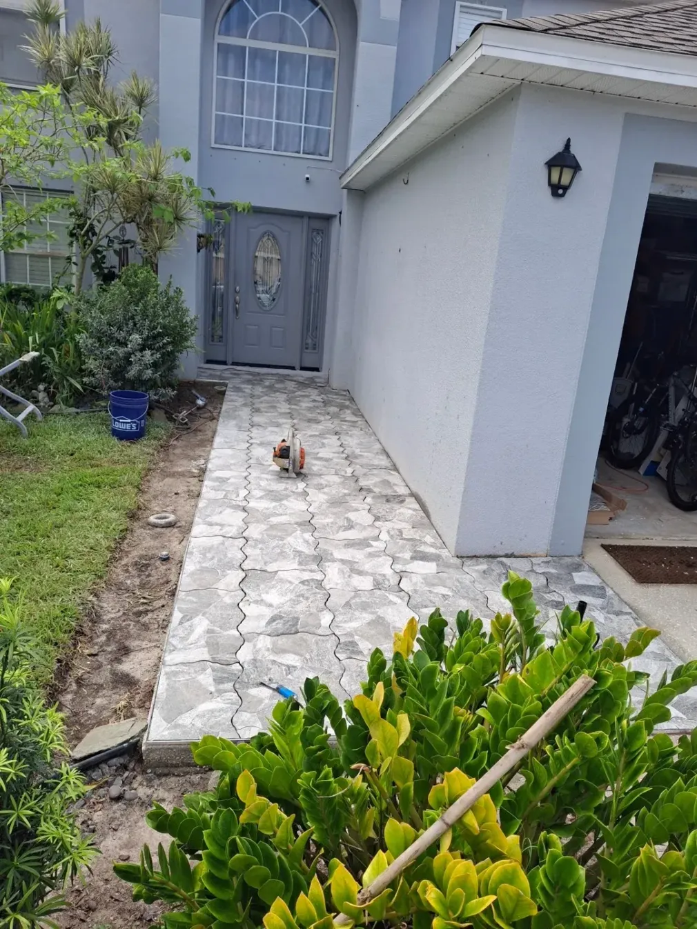 A newly paved gray stone walkway leads to the front door of a suburban house, flanked by green bushes and a lawn.