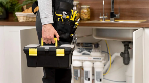 A repair person holding a toolbox stands in front of an open kitchen cabinet containing a water filtration system.