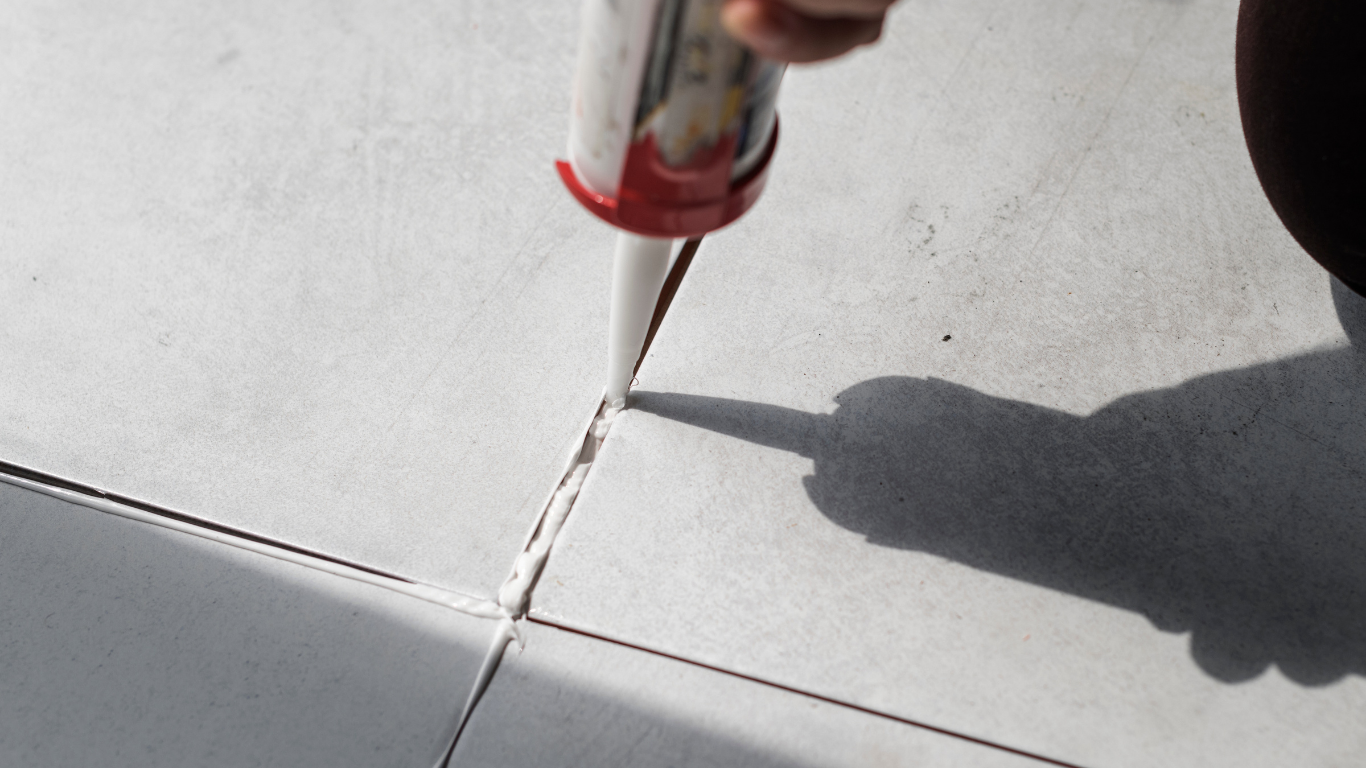 A hand uses a caulking gun to apply sealant into the gap between gray ceramic floor tiles.