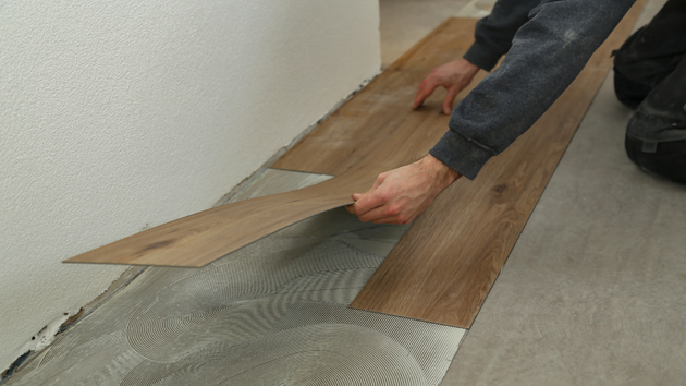 A person installing wood-look vinyl flooring over a thin layer of adhesive on a subfloor next to a white wall.