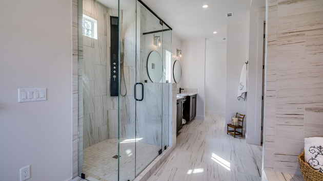 A modern bathroom featuring a glass shower, marble walls, white flooring, a dark vanity, and a small wooden stool.