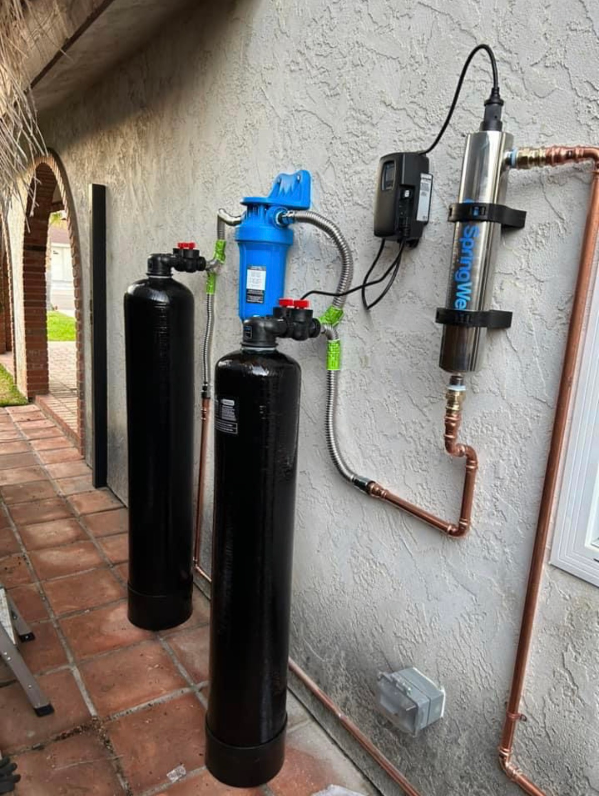 Water filtration system with black tanks and blue pump mounted on an exterior wall.