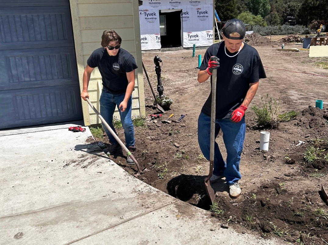 Two workers shoveling dirt beside a concrete walkway near a house with a garage door.
