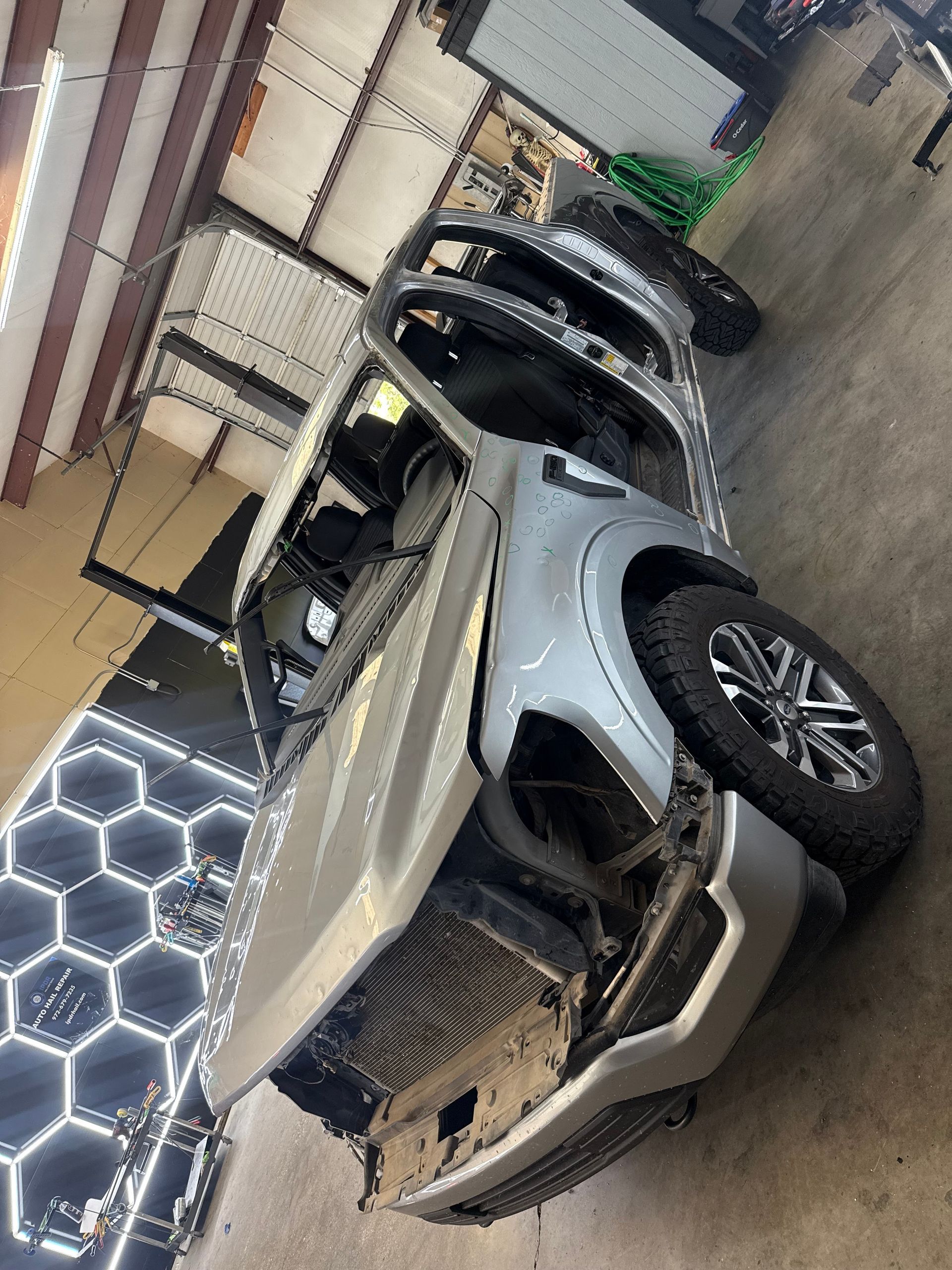 Damaged silver pickup truck in a shop, front end crumpled, driver's side damaged.